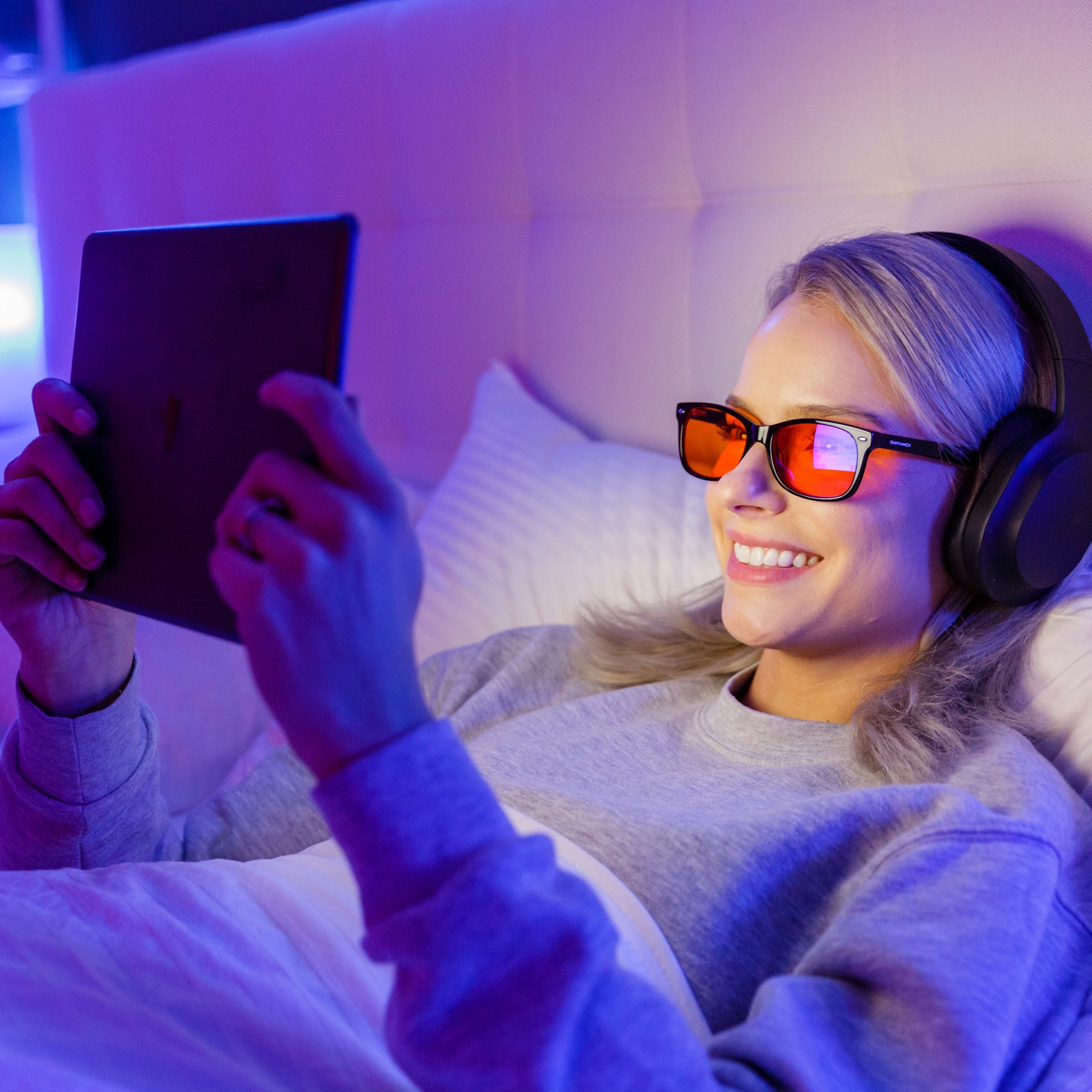 Person lying on a bed using a tablet with headphones and blue-light blocking glasses in a dimly lit room.