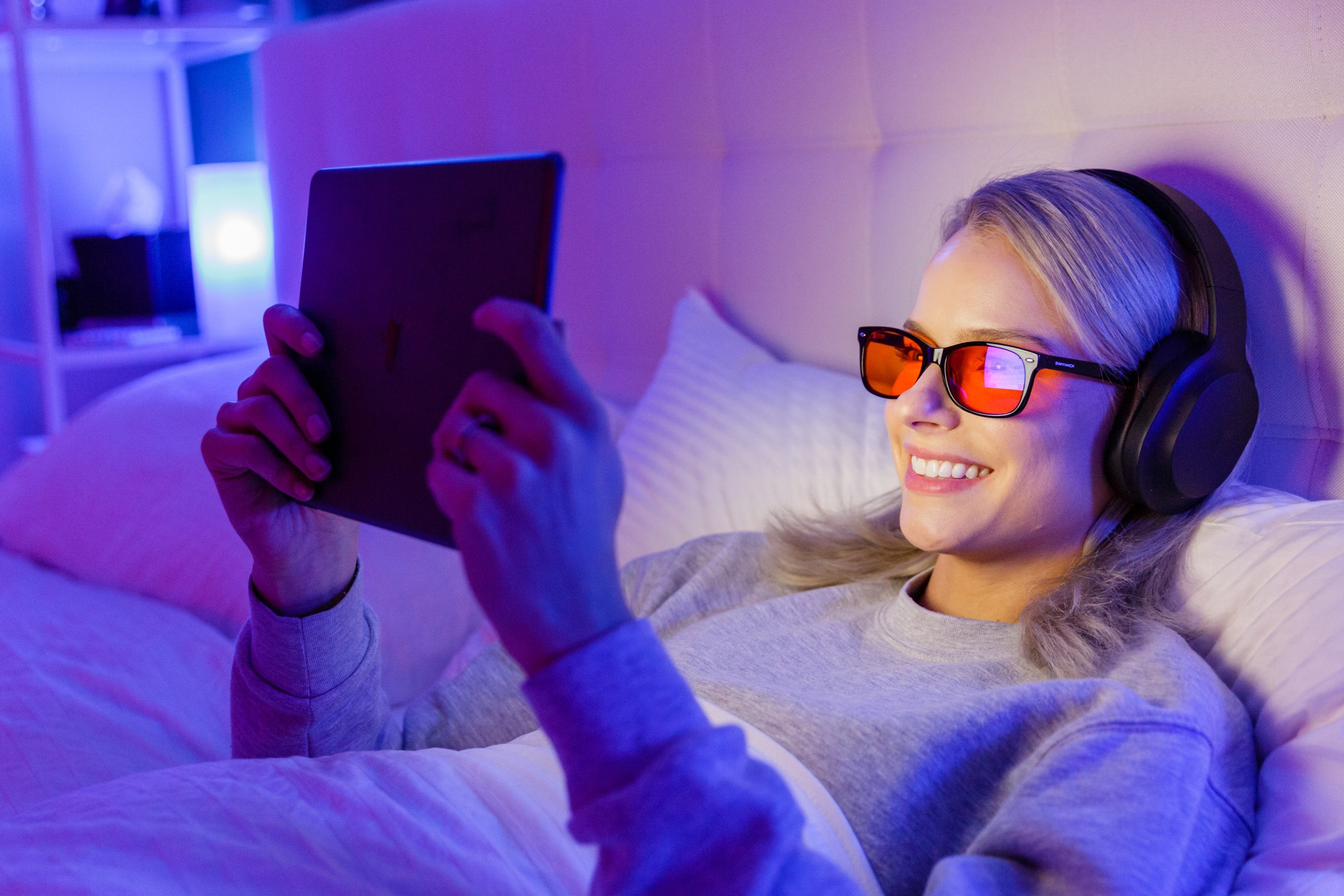 Person lying on a bed using a tablet with headphones and blue light blocking glasses in a dimly lit room.