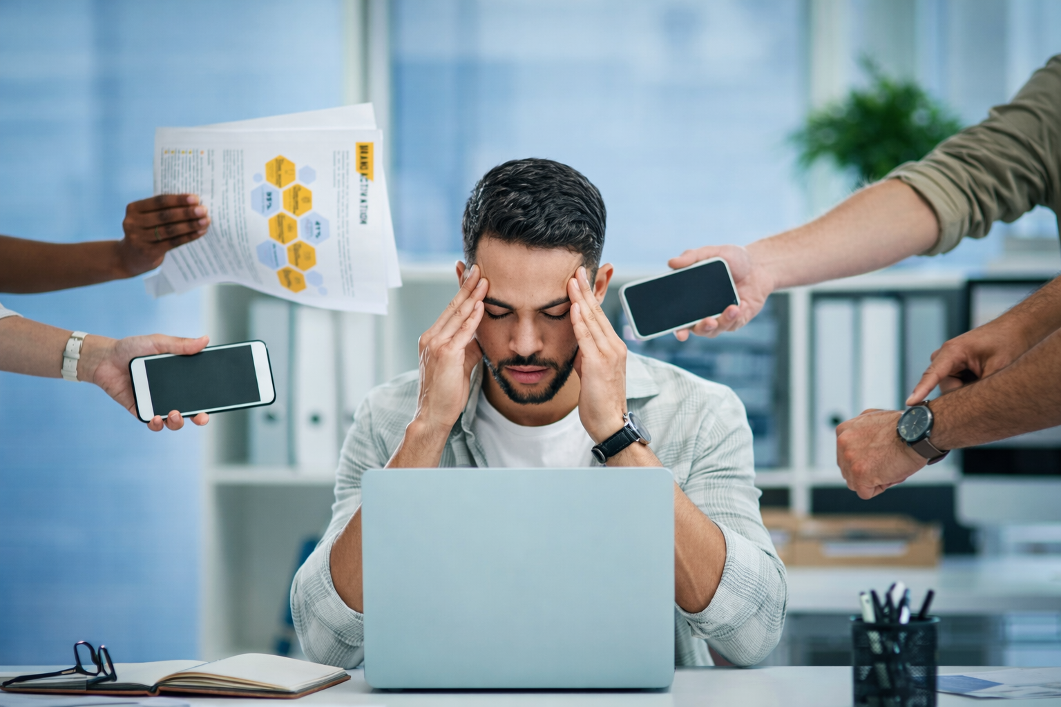 Stressed man at a desk with multiple people around him holding smartphones and documents.