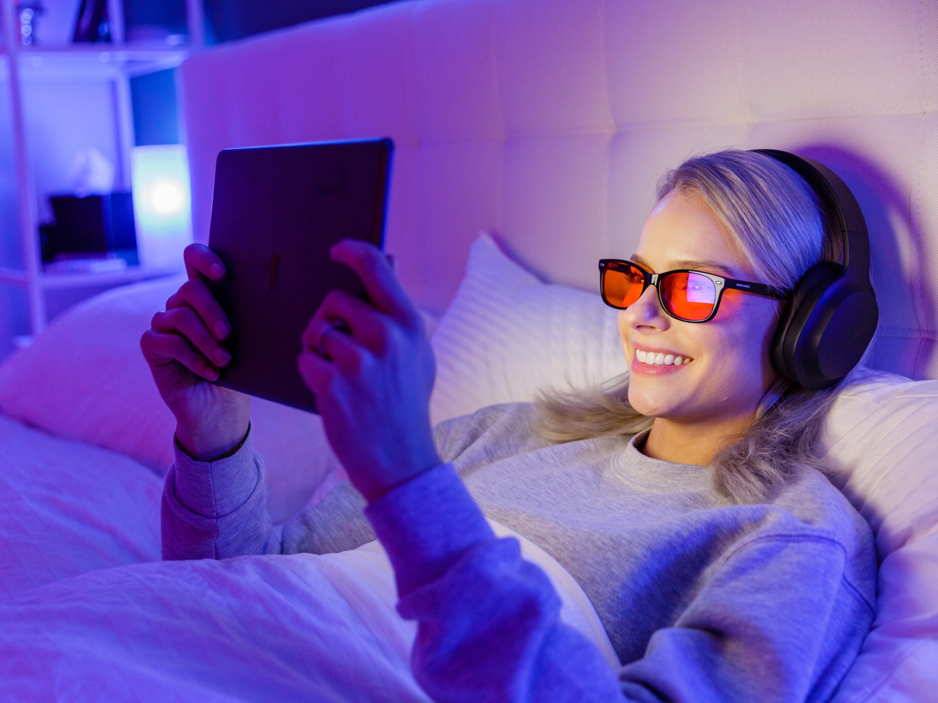 Person lying on a bed using a tablet with headphones and blue-light blocking glasses in a dimly lit room.