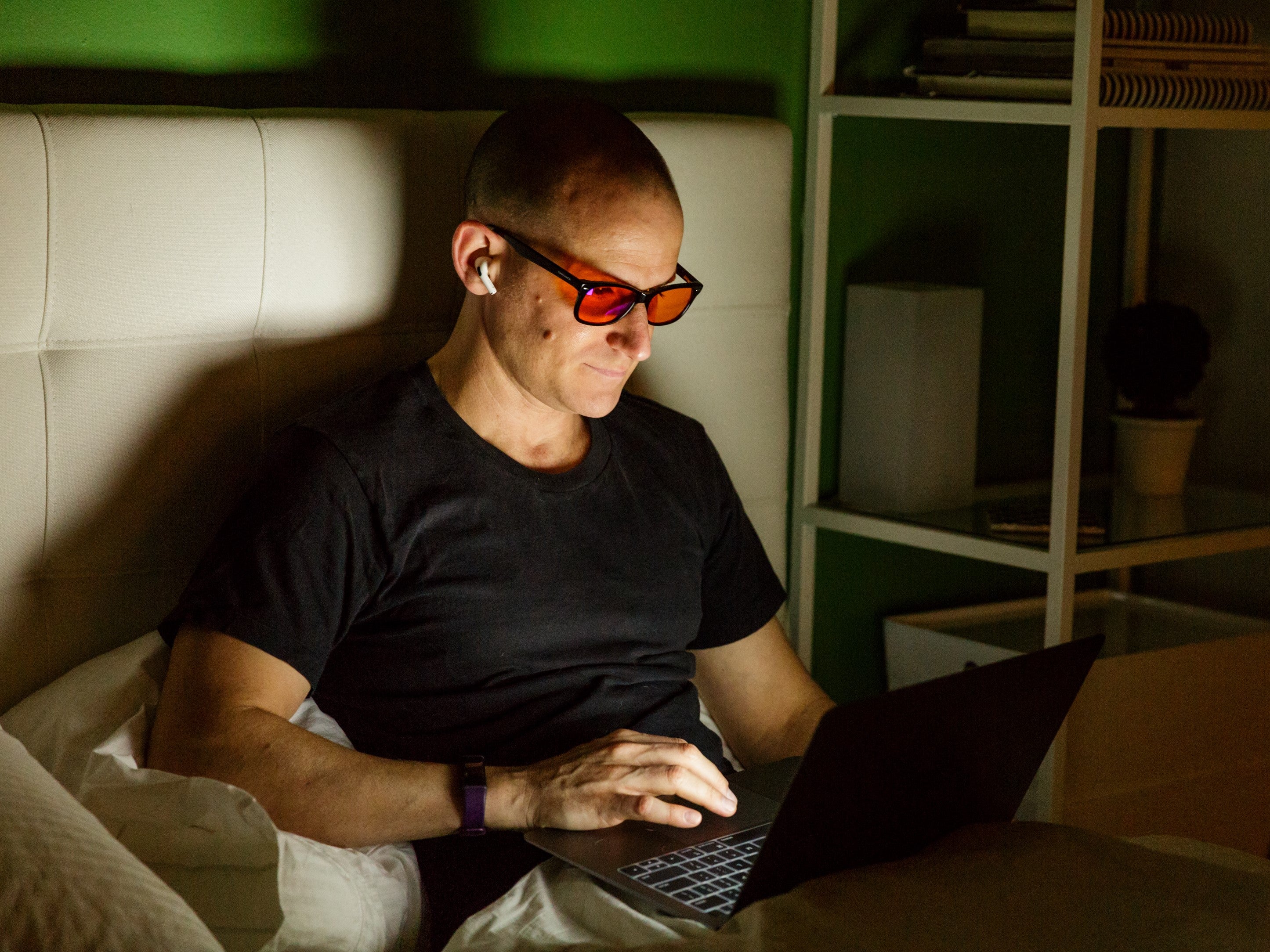 Man wearing blue light blocking glasses using a laptop in a dimly lit room.